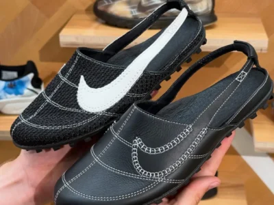 Hands hold two Nike Moon Shoe Slingback prototypes inside a sneaker showroom. The black backless shoes feature a vintage Moon Shoe-inspired upper with exposed white contrast stitching, waffle-inspired outsole lugs, and slingback heel straps. One pair is finished in smooth black leather with a stitched Swoosh, while the other uses textured black material with a large white leather Swoosh extending onto the heel strap. Wooden display shelves filled with Nike footwear appear softly blurred in the background