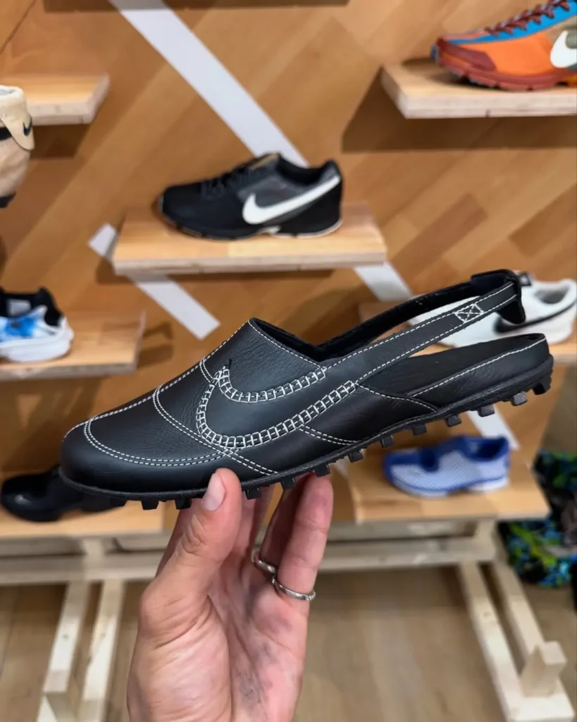 A hand holds a black Nike Moon Shoe Slingback in profile inside a sneaker boutique. The backless design combines premium black leather, oversized white contrast stitching tracing the Swoosh and panel lines, an adjustable slingback strap, and a rugged waffle-style outsole inspired by Nike's original Moon Shoe. Wooden shelving and classic Nike sneakers create a warm retail backdrop