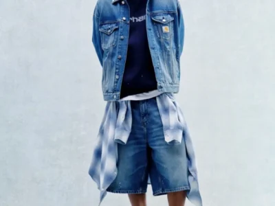 Model wears a light-wash denim trucker jacket over a navy Carhartt WIP sweatshirt, oversized denim shorts, and tan suede clogs, with a blue plaid shirt tied around the waist