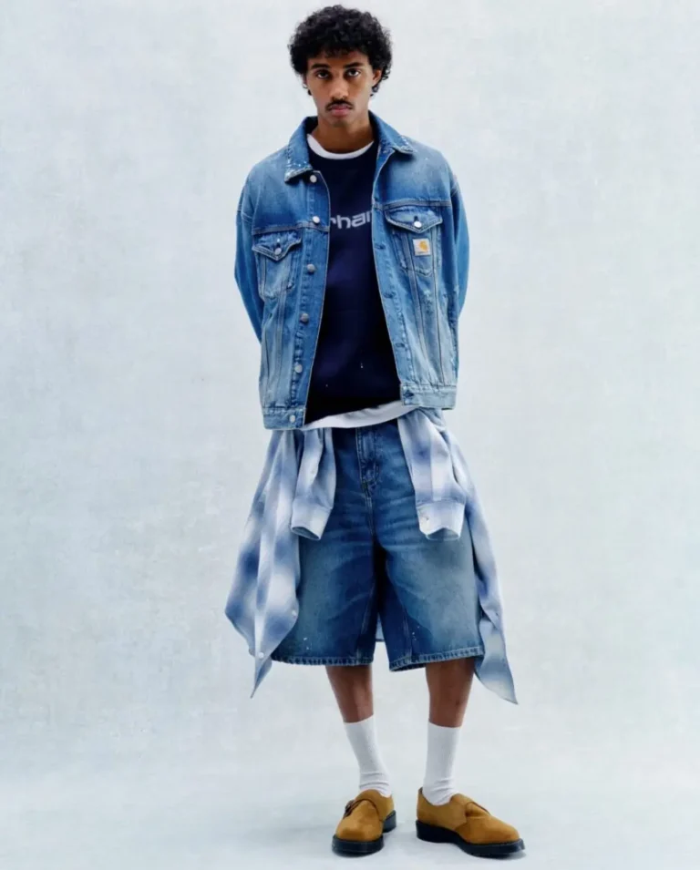 Model wears a light-wash denim trucker jacket over a navy Carhartt WIP sweatshirt, oversized denim shorts, and tan suede clogs, with a blue plaid shirt tied around the waist