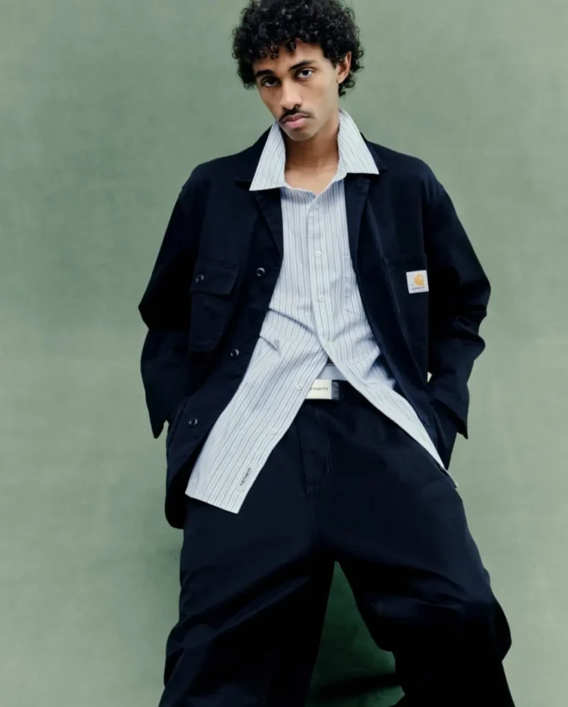 Model poses in a navy utility suit featuring a workwear-inspired jacket and wide-leg trousers, layered over a striped button-down shirt from the Carhartt WIP FW26 collection
