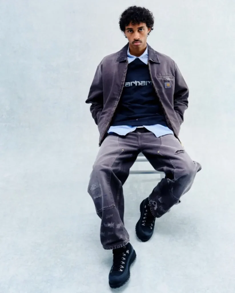 Model seated wearing a faded charcoal work jacket and matching distressed carpenter pants, styled with a navy logo sweatshirt, light blue shirt, and rugged black boot