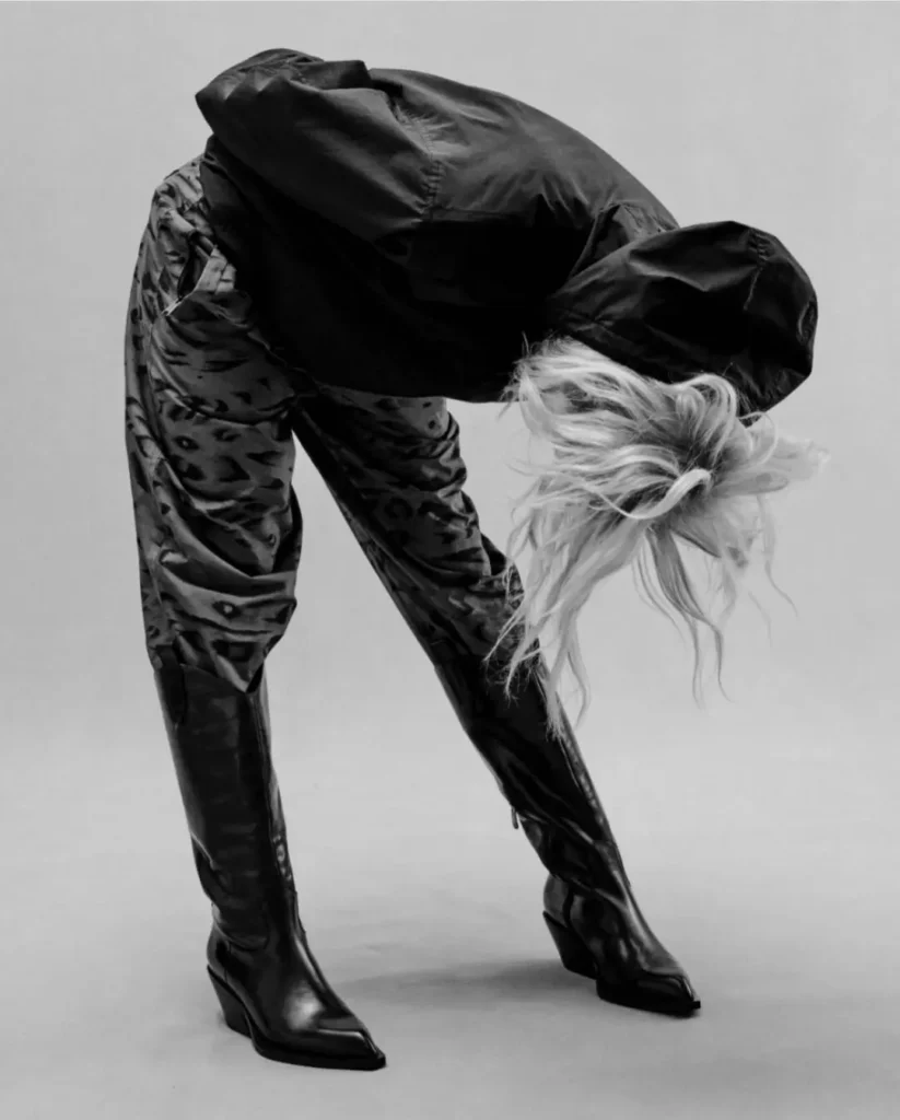 Black-and-white editorial image of a model bent forward wearing a black oversized jacket and patterned thigh-high boots, highlighting the collection’s experimental fashion direction