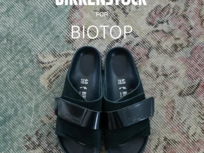 Birkenstock for Biotop black slide sandals displayed on a vintage-patterned rug, showcasing minimalist crossover straps and tonal monochrome styl