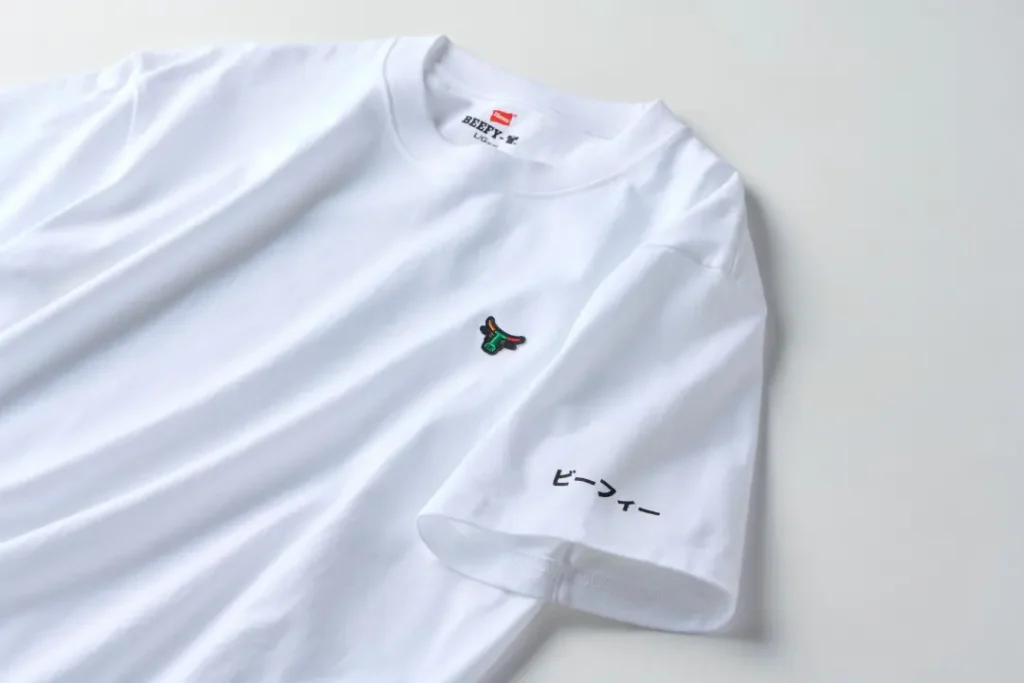 Angled close-up of the white Hanes BEEFY T-shirt highlighting the embroidered bull-head chest logo and Japanese sleeve graphic. Soft folds emphasize the heavyweight cotton fabric and relaxed fit.
