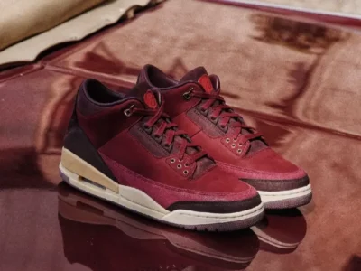 Pair of Air Jordan 3 Low BIN23 shoe displayed on a reflective burgundy surface, featuring premium wine-red leather and suede construction with aged cream midsoles and refined luxury detail