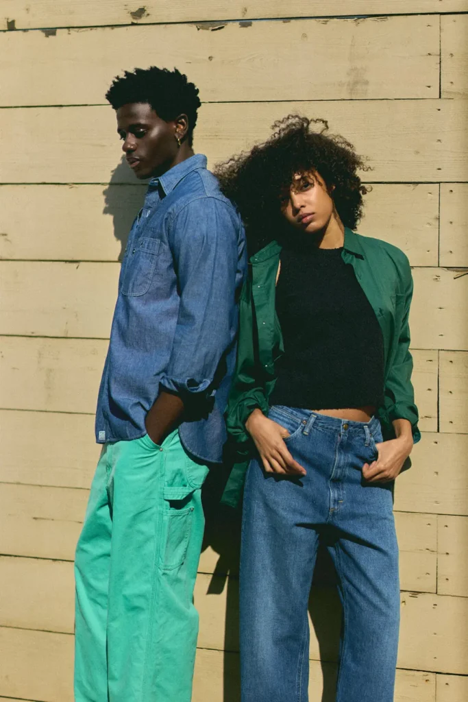 A male and female model lean against a sunlit wooden backdrop, showcasing casual denim and utility-inspired separates with a laid-back contemporary aesthetic