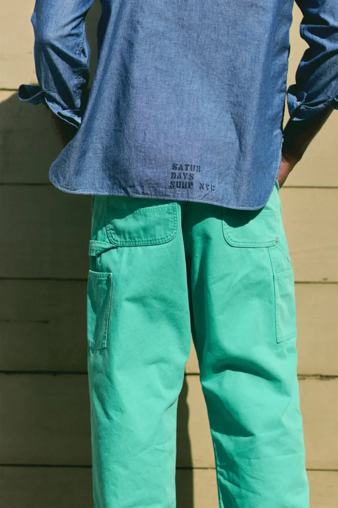 Close-up rear view of mint-green carpenter pants paired with a light indigo chambray shirt, highlighting oversized utility pockets and relaxed tailor