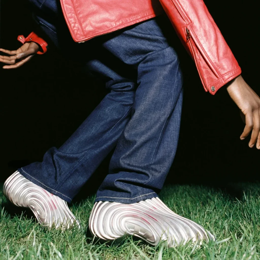 Close-up of a model wearing metallic silver Melissa × Diesel clogs beneath dark denim trousers, emphasizing the futuristic molded sole and reflective texture
