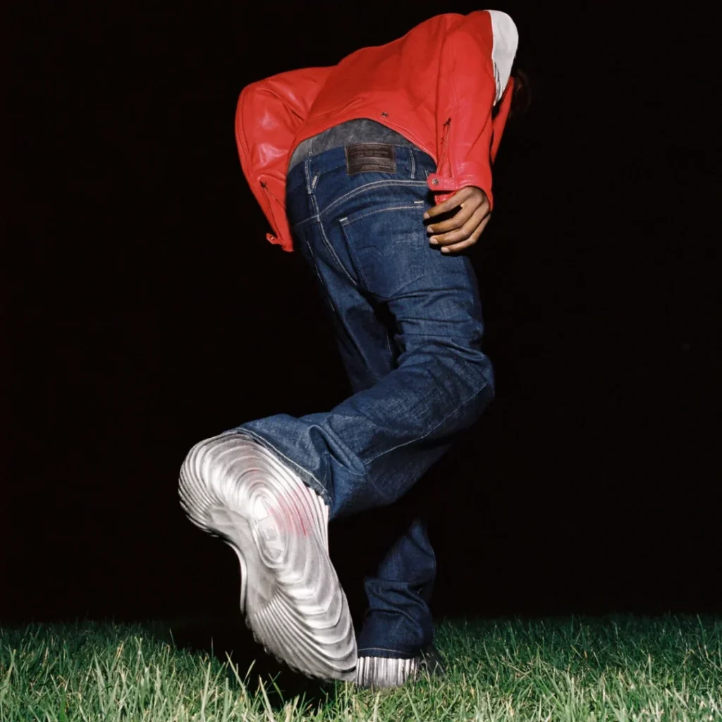 Campaign image of a model in a red leather jacket and denim jeans walking across grass, featuring oversized metallic silver Melissa × Diesel clogs with sculpted wave detail