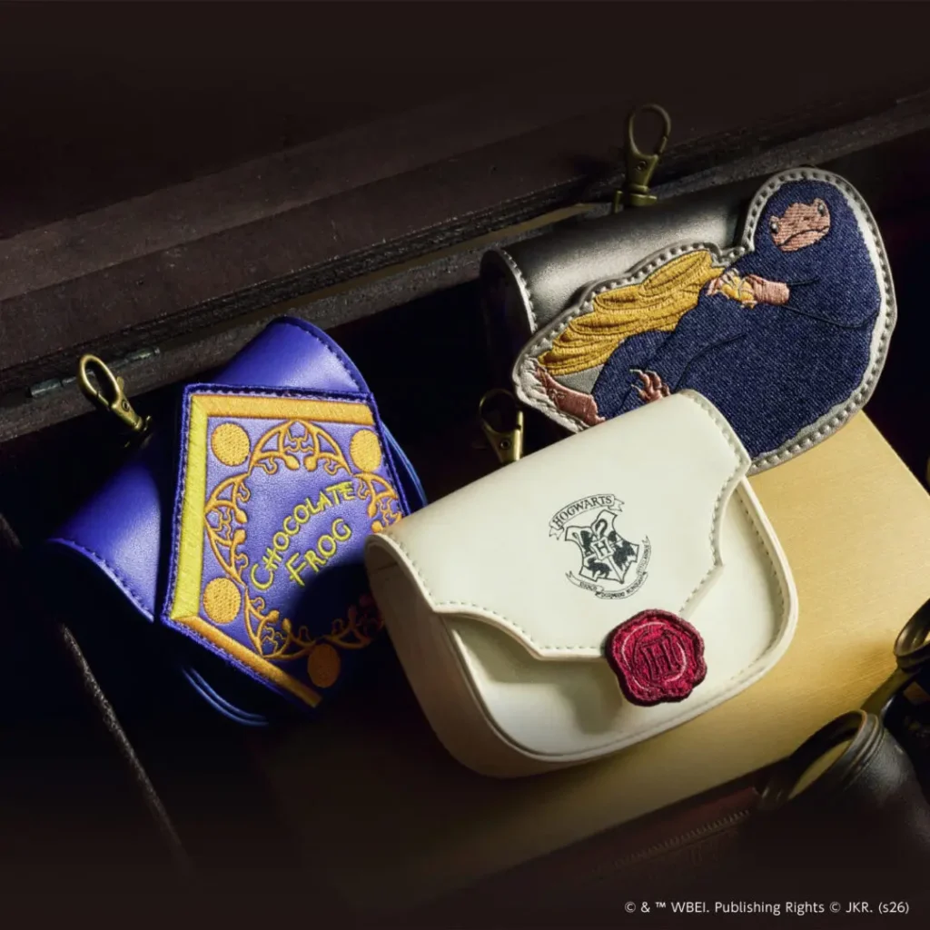 Close-up of Harry Potter-themed accessories displayed inside a vintage-style trunk, including a white Hogwarts envelope pouch with a red wax-seal detail, a purple Chocolate Frog-inspired coin pouch, and an embroidered Niffler character pouch with gold coin accent