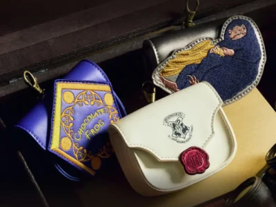 Close-up of Harry Potter-themed accessories displayed inside a vintage-style trunk, including a white Hogwarts envelope pouch with a red wax-seal detail, a purple Chocolate Frog-inspired coin pouch, and an embroidered Niffler character pouch with gold coin accent
