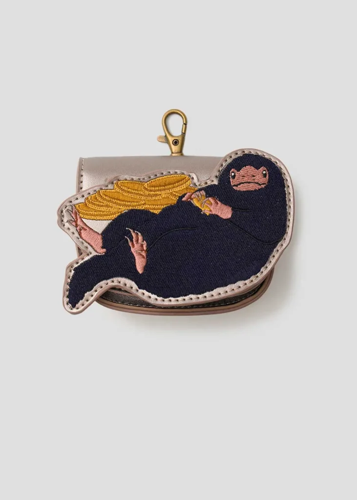 Niffler-inspired coin pouch with embroidered character artwork holding gold coins, attached to a taupe leather-look pouch with a brass-tone clasp and soft neutral backdrop