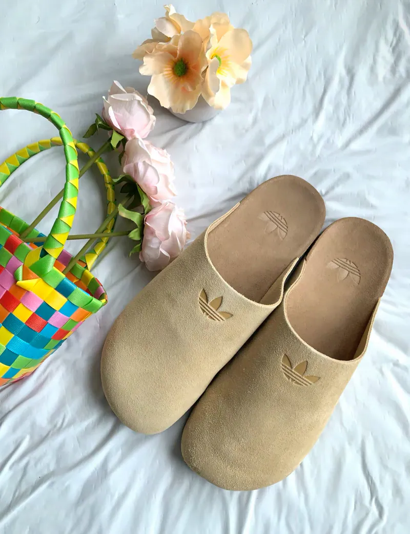 Beige adidas mule slippers arranged on fabric alongside flowers and a colorful woven basket for a lifestyle present