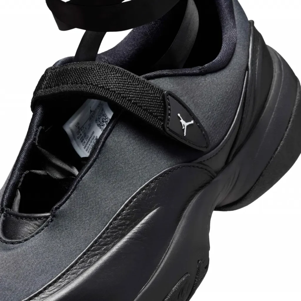 Close-up detail of the Air Jordan shoe showing the textured strap closure, glossy Jumpman badge, and premium black leather and textile material