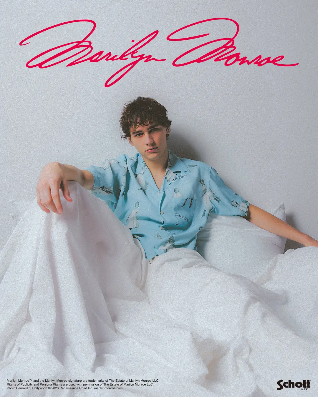 A model lounges across crisp white bedding while wearing a light blue short-sleeve camp-collar shirt covered in vintage Marilyn Monroe illustrations and signature motifs. The relaxed styling, soft pastel palette, and minimalist bedroom setting create a nostalgic Hollywood-inspired mood, highlighting the shirt’s retro graphics and breezy summer sil