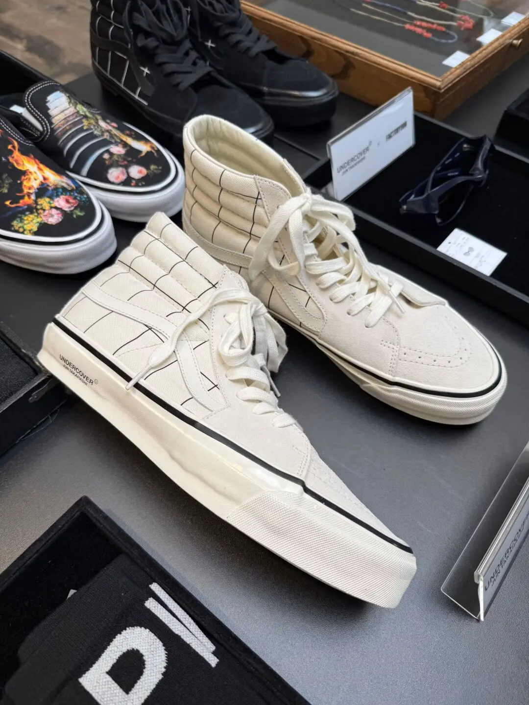 Cream-colored UNDERCOVER x Vans Sk8-Hi shoe with subtle black grid graphics and oversized padded collars. The minimalist design contrasts with the darker styles displayed alongside them in the showroom present