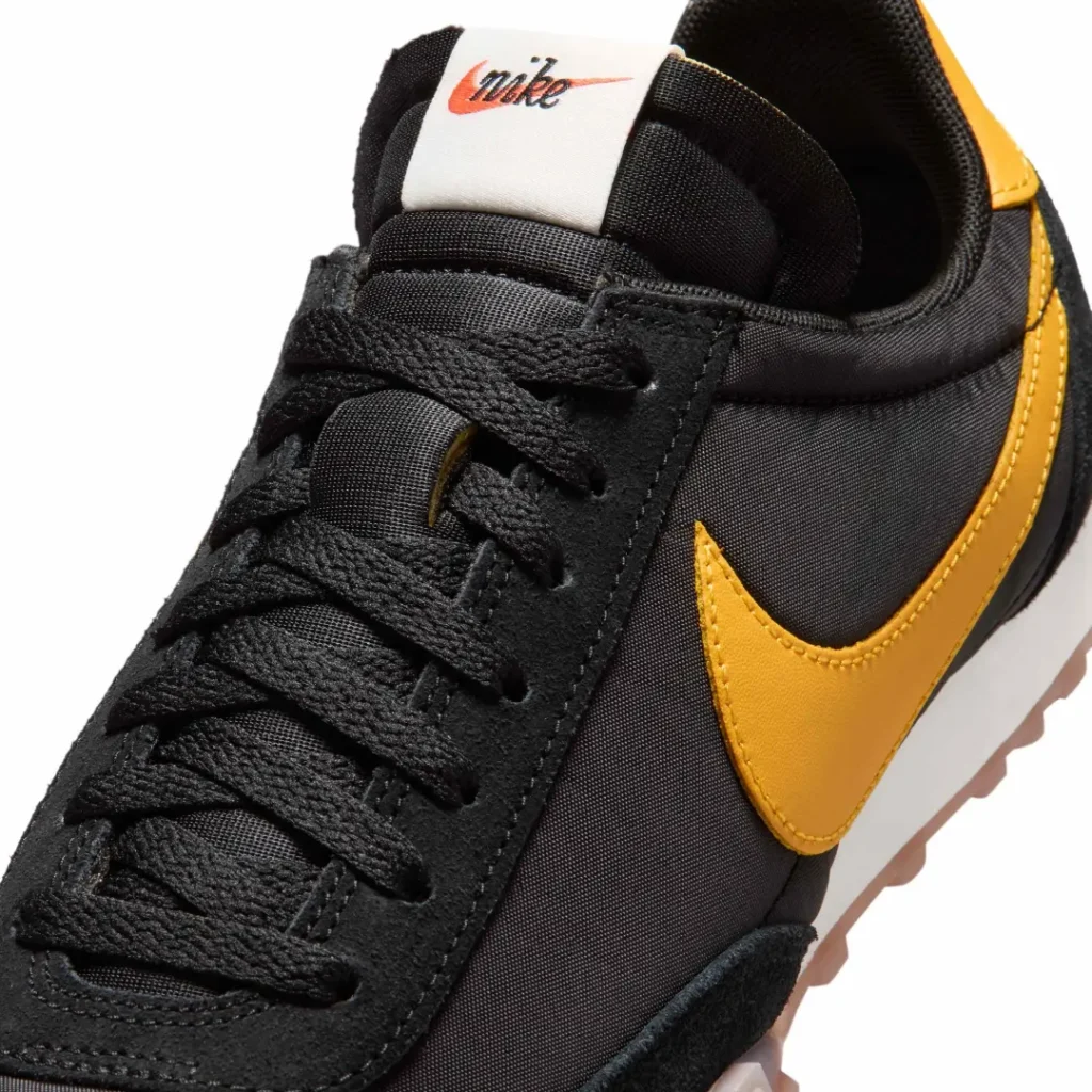 Close-up of the Nike sneaker’s forefoot and midfoot, emphasizing textured black suede overlays, nylon materials, and a bold yellow Swoosh