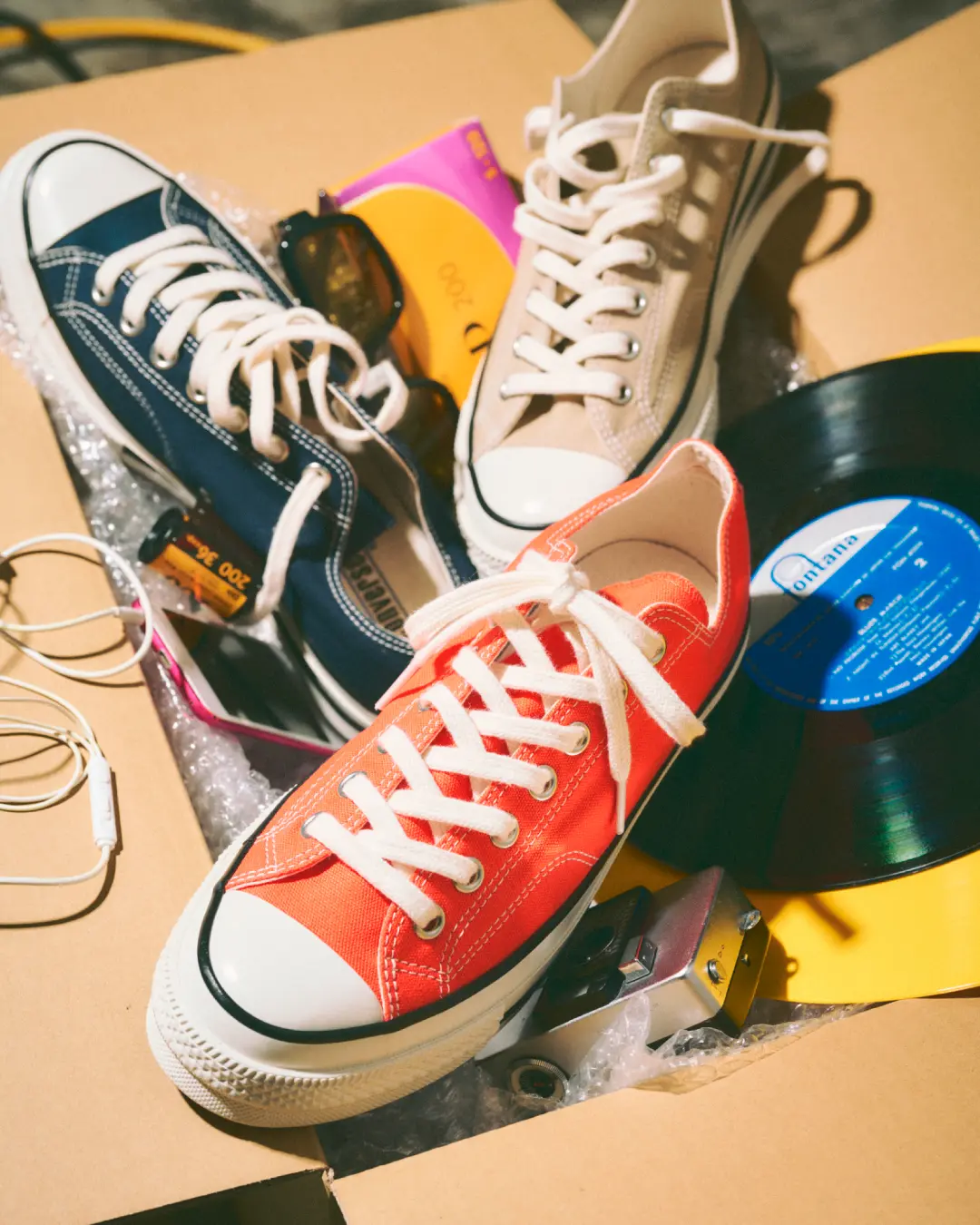 Converse Chuck Taylor All Star low-top sneakers in vibrant orange, navy, and beige colorways arranged among vinyl records, film cameras, sunglasses, earphones, and retro accessories inside an open cardboard box