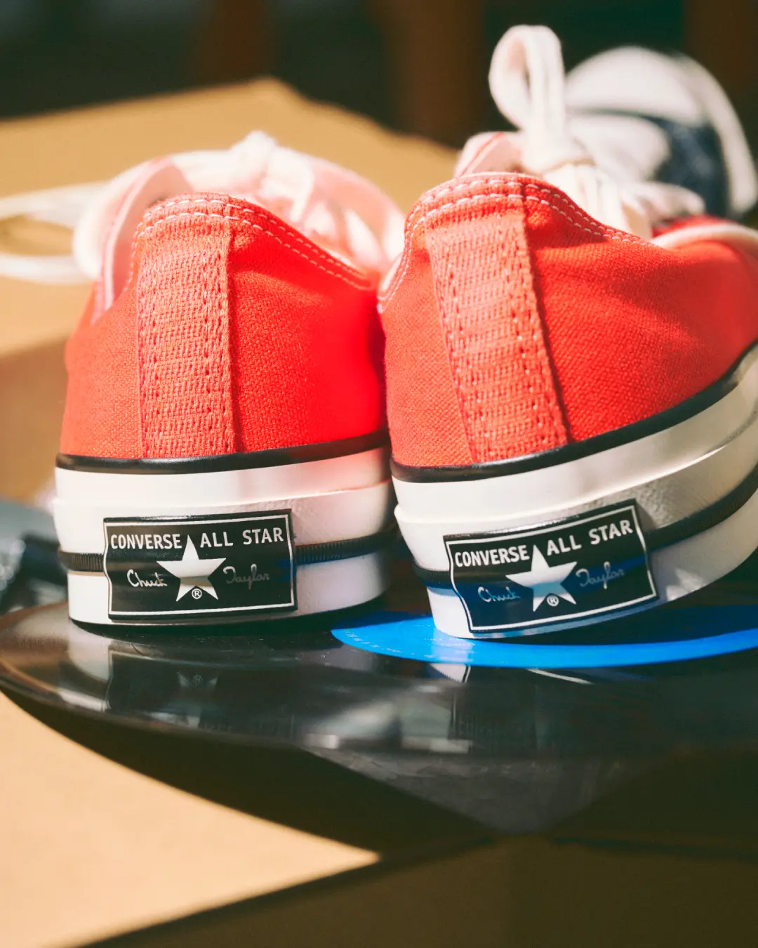 Rear view of bright orange Converse Chuck Taylor All Star low-top shoe resting on a vinyl record, showcasing the iconic black-and-white heel license plate brand