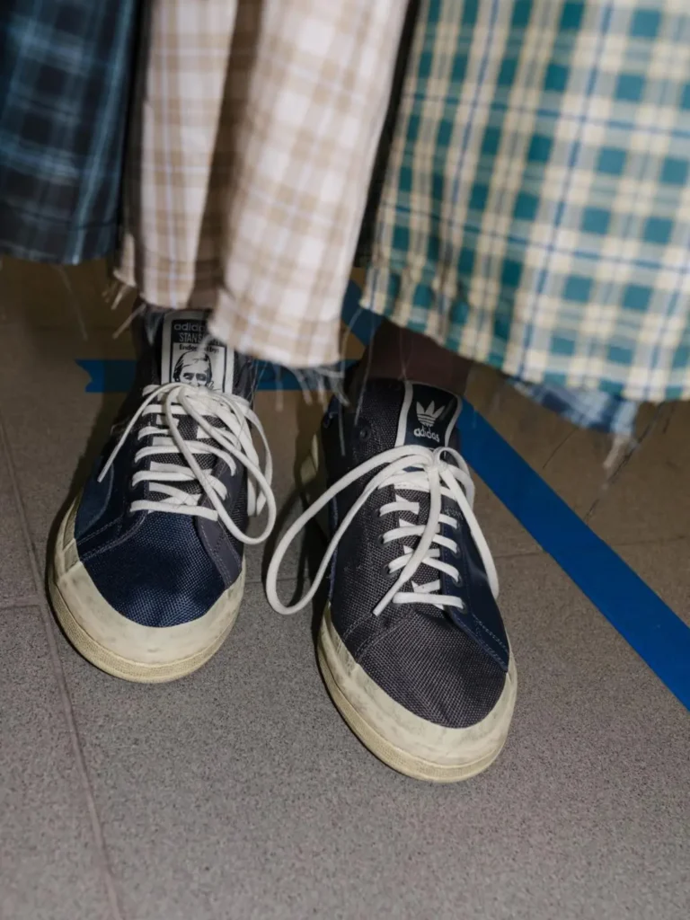 Close-up of navy adidas Stan Smith sneakers worn beneath layered plaid garments, revealing aged midsoles and long white lace