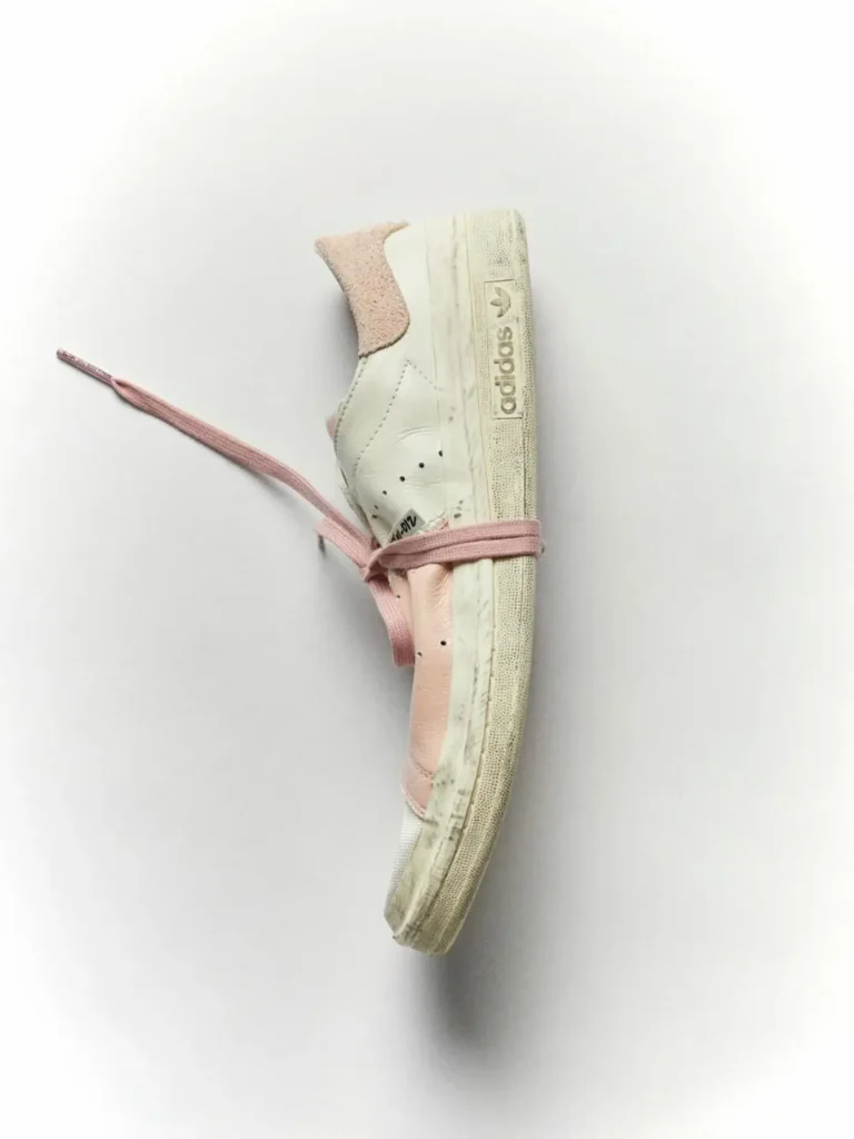 Side profile of a white and blush pink adidas Stan Smith shoe suspended against a white background, featuring weathered foxing and pink lace