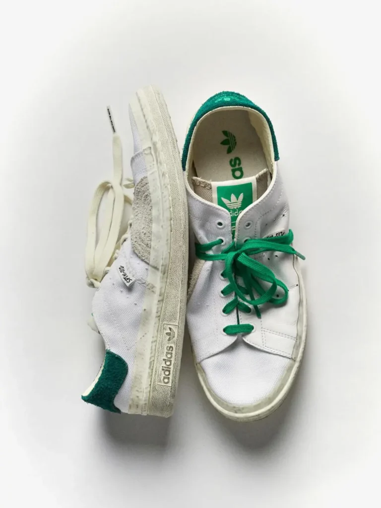 White adidas Stan Smith shoe with green accents suspended against a white background, highlighting distressed canvas uppers and green lace