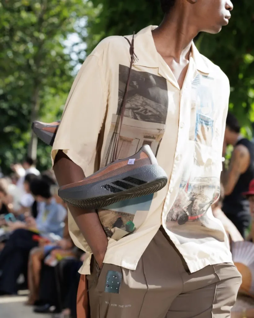 Model wears a printed short-sleeve shirt while carrying a streamlined adidas shoe as a shoulder bag during an outdoor runway presentation