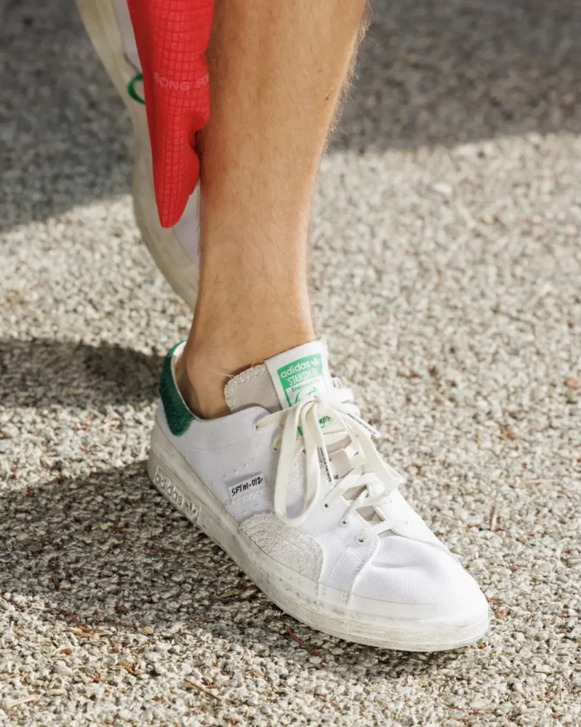 lose-up of a white adidas Stan Smith shoe with green heel tab worn on a gravel runway, highlighting distressed canvas and textured overlay