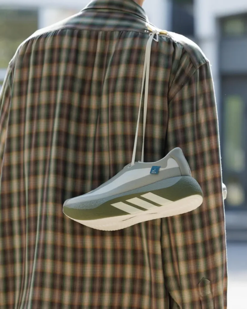 Olive and gray adidas shoe hangs from the back of a plaid shirt by its lace, styled as a shoulder accessory