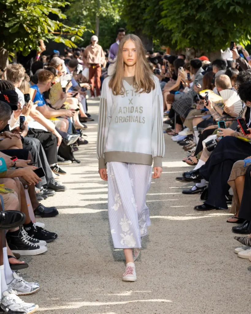 Runway model wears a light gray adidas Originals sweatshirt with a floral skirt and blush pink adidas Stan Smith shoe during the outdoor show