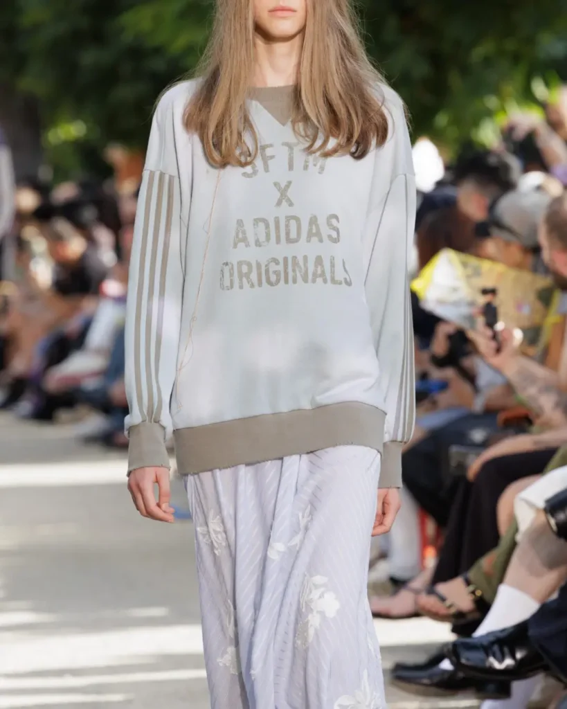Close-up of a runway look featuring a faded gray adidas Originals x Song for the Mute sweatshirt with three-stripe sleeves, paired with a flowing floral skirt