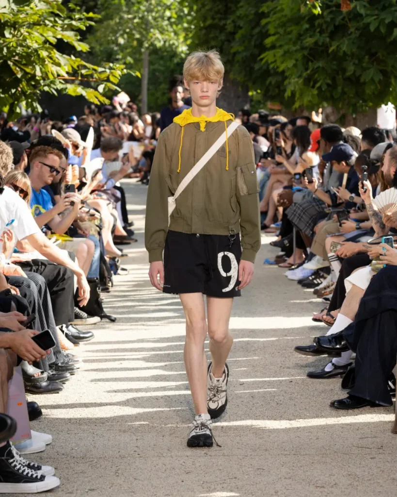Model walks an outdoor runway wearing an olive hooded jacket, black shorts with a large numeral graphic, crossbody bag, and adidas trail-inspired shoe