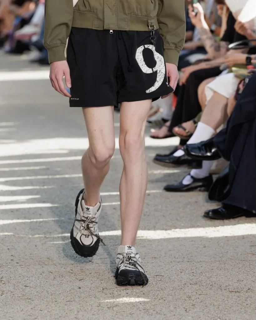 Close-up of black graphic shorts and adidas technical trail sneakers with chunky soles, highlighting the footwear and lower-half style