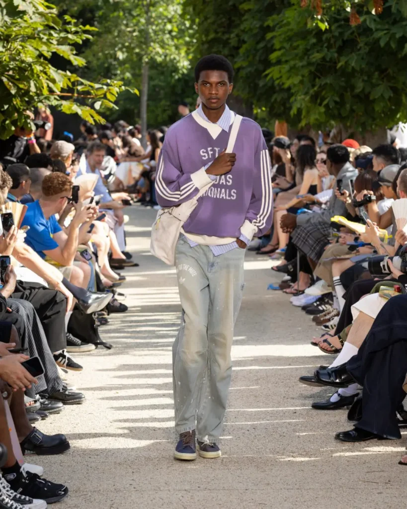 Runway model wears a lavender adidas Originals x Song for the Mute sweatshirt layered over a striped shirt with faded denim, navy Stan Smith shoes, and a white shoulder bag