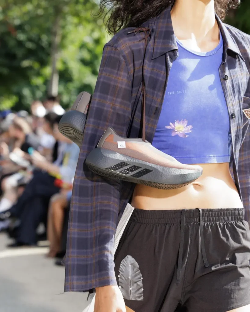 Close-up of a blue floral crop top beneath a navy plaid shirt, paired with black shorts and a streamlined adidas shoe worn as a crossbody accessory
