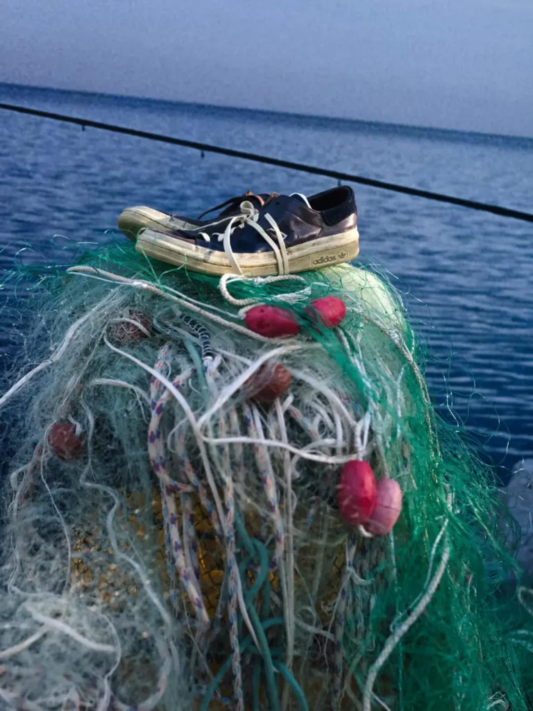 Worn navy Adidas shoe rest atop bundled green fishing nets and red floats beside calm blue water, highlighting weathered materials and coastal inspiration