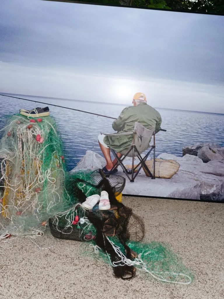Installation of fishing nets, baskets, and sneakers sits before a large waterside photograph featuring an angler, emphasizing the collection's maritime storytell