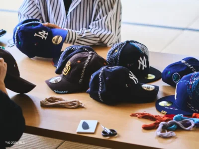A behind-the-scenes workshop table displays several customized New Era MLB caps, including New York Yankees, Los Angeles Dodgers, and San Diego Padres designs. The hats are decorated with colorful hand-stitched embroidery, while embroidery thread, scissors, and crafting materials are scattered across the wooden surface as designers work in the background