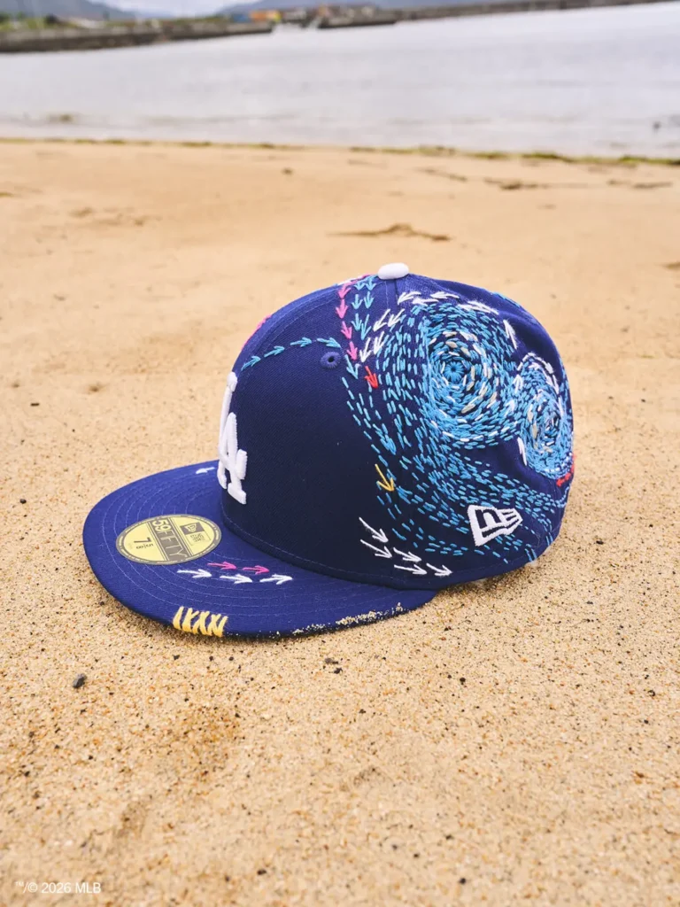 A Los Angeles Dodgers New Era fitted cap rests on beach sand near the shoreline. The royal blue cap is decorated with swirling blue embroidery, colorful stitched arrows, and handcrafted details that extend across the crown and visor, blending sportswear with artisanal design