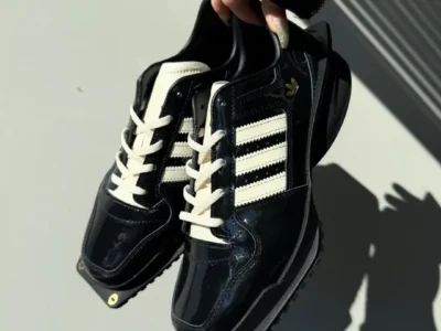 Hand holding a pair of glossy black adidas Forum shoe featuring cream Three Stripes, off-white laces, gold Trefoil branding, and an exaggerated sculpted sole under bright natural light