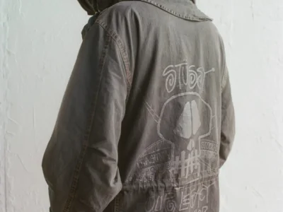 Back view of a faded charcoal hooded jacket featuring hand-drawn Stüssy and Our Legacy Workshop graphics across the rear panel, styled with a washed, worn-in finish against a minimalist studio backdrop