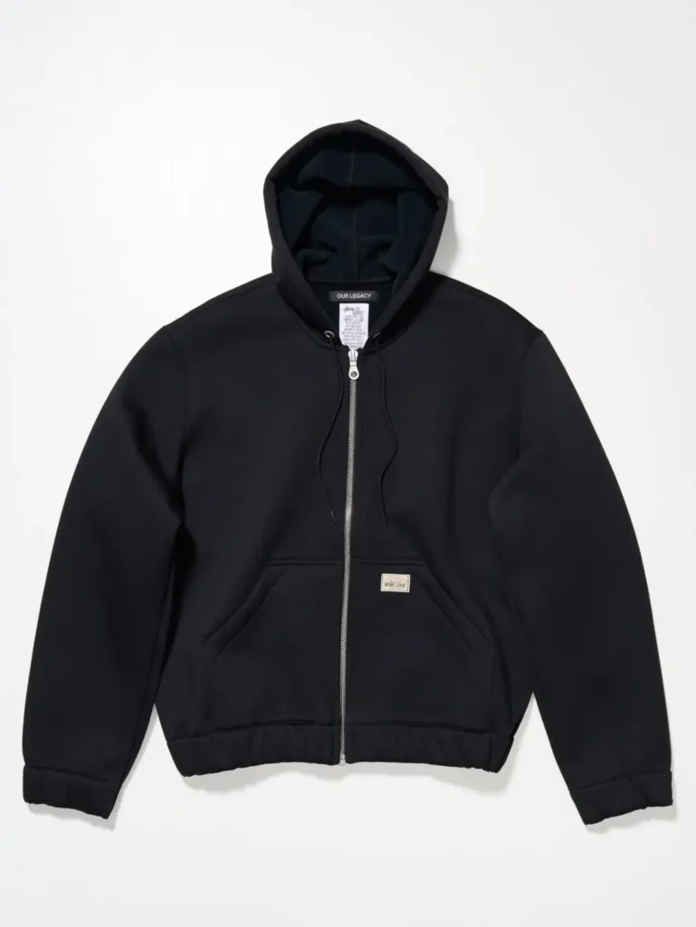 Front view of a black zip-up hoodie with clean minimalist construction, silver hardware, and collaborative Stüssy Our Legacy Workshop label detai