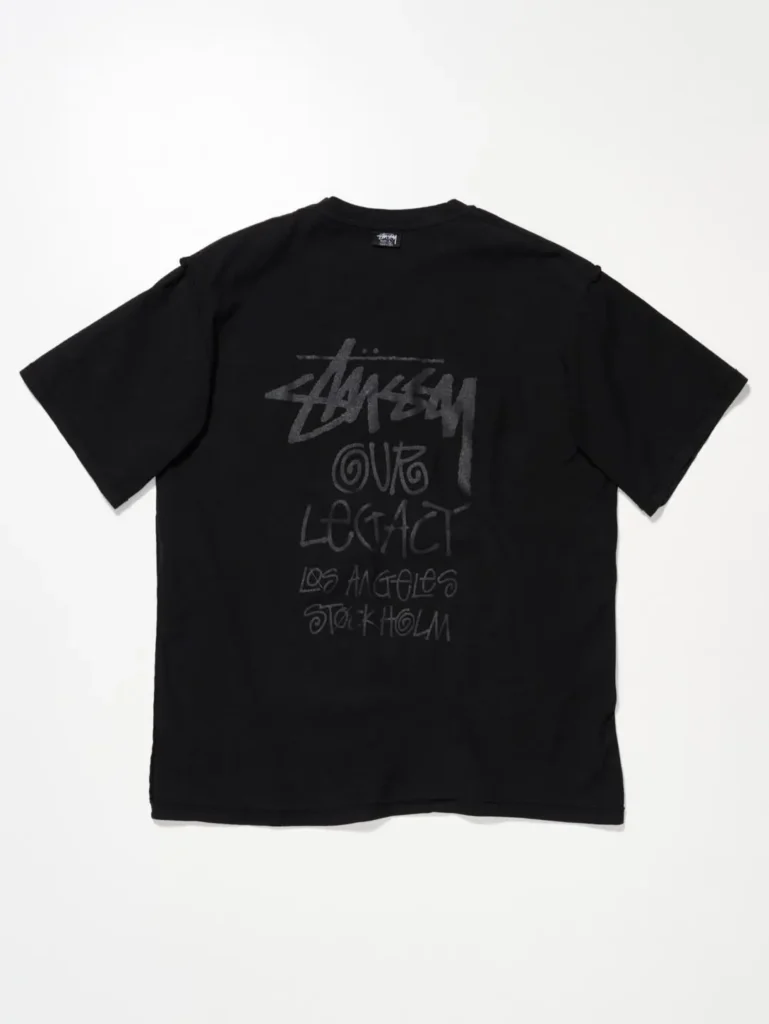 Black Tee with tonal back graphic displaying handwritten-style Stüssy branding alongside collaborative workshop-inspired typography
