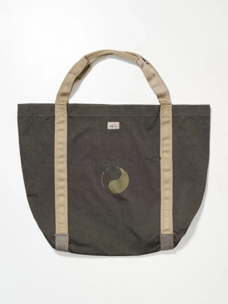 Large tote bag in muted brown-grey fabric with contrasting beige handles and subtle collaborative branding centered on the exterior