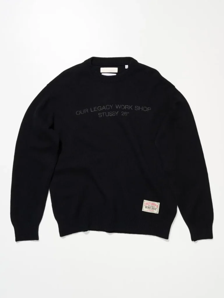 Black crewneck sweater with "OUR LEGACY WORK SHOP / STUSSY 26"" embroidered in tonal black text across the chest, plus a woven Stüssy Garments x Our Legacy Work Shop label near the hem