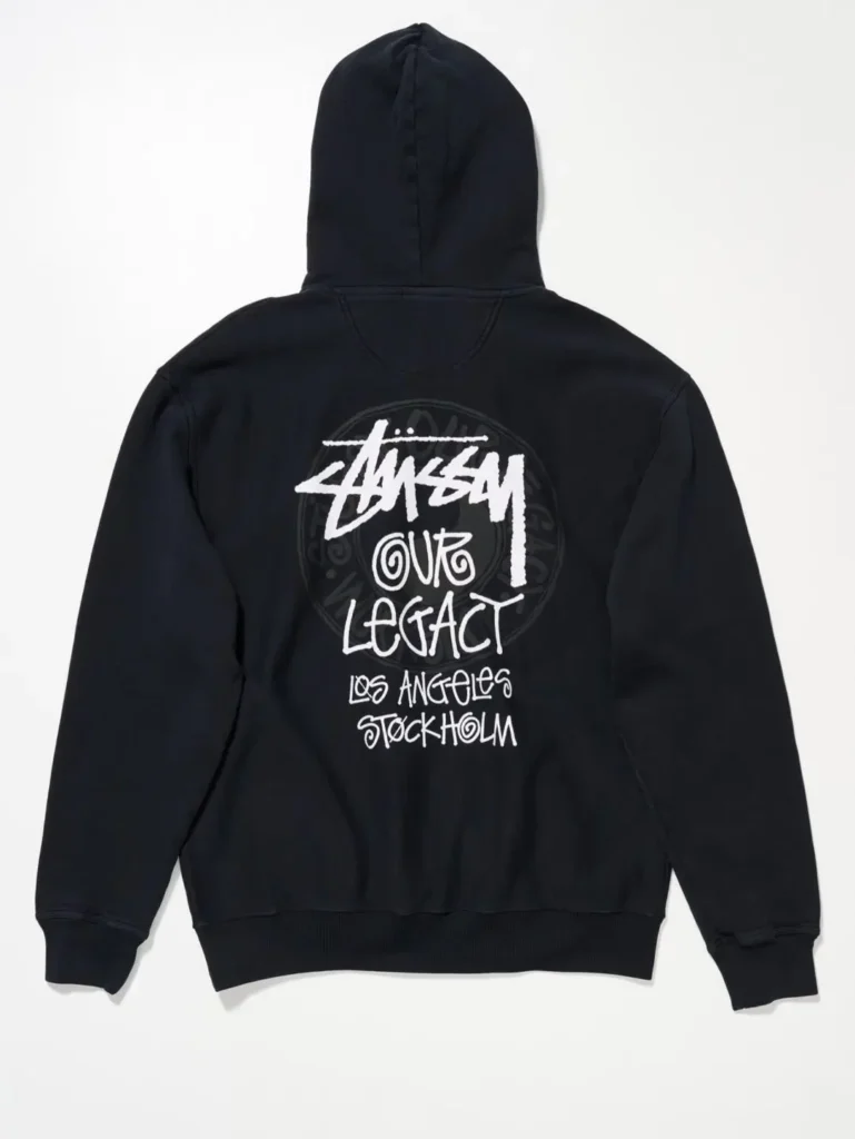Black pullover hoodie, back view, printed with a large white Stüssy-style graffiti logo, "Our Legacy" script, "Los Angeles / Stockholm" text, and a faint tonal circular stamp graphic behind the letter
