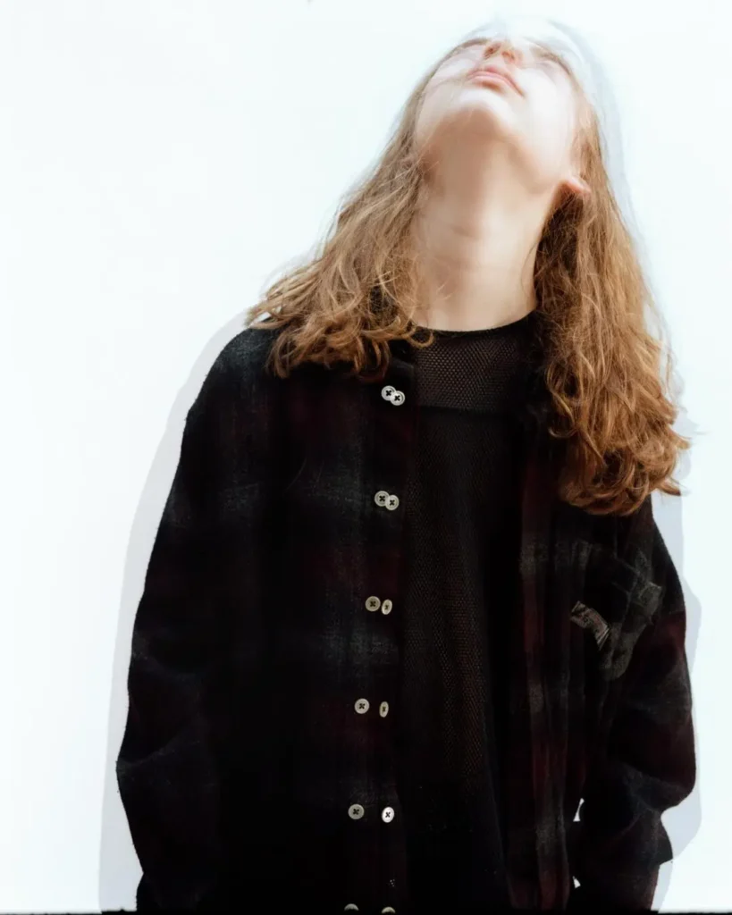 Model wearing a dark plaid overshirt over a sheer black mesh top, photographed against a bright white background with soft motion blur creating an ethereal editorial effect