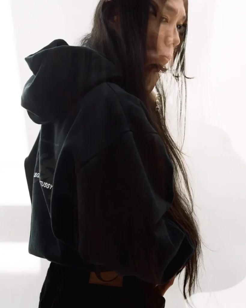 Editorial image of a model in a cropped black hoodie featuring subtle collaborative branding, captured with layered exposure and soft natural light