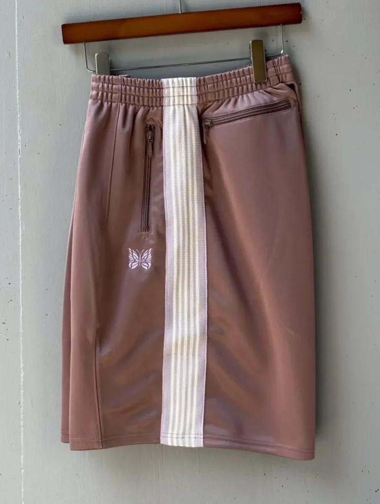 Dusty pink Needles track shorts hanging on a wooden hanger with a white striped center panel, zip pockets, elastic waistband, and a white butterfly logo, highlighting the exclusive colorway and premium finish
